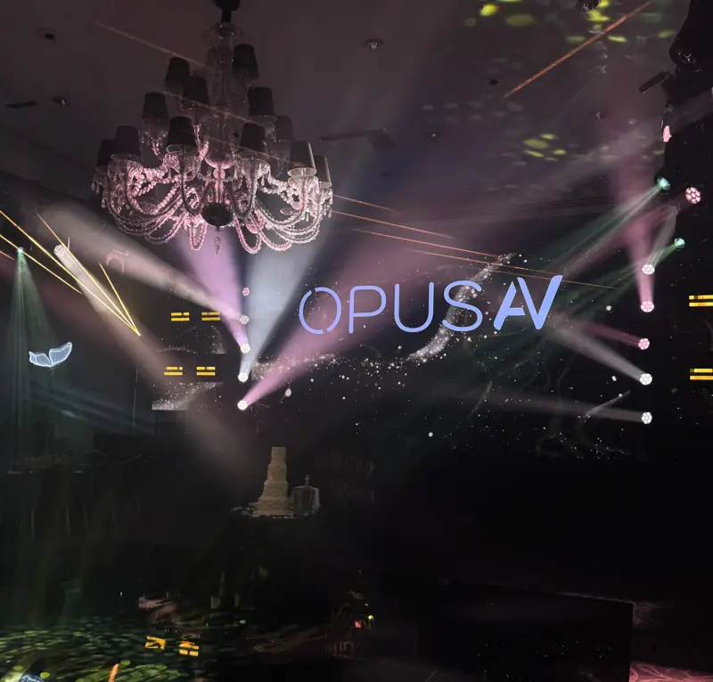 Ballroom lit with moving-head beams under a chandelier, the Opus AV logo projected on the wall